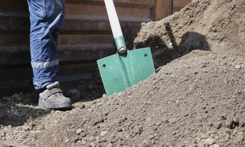 A close up shot of a shovel in dug out sand and the yard leveling professional standing beside it
